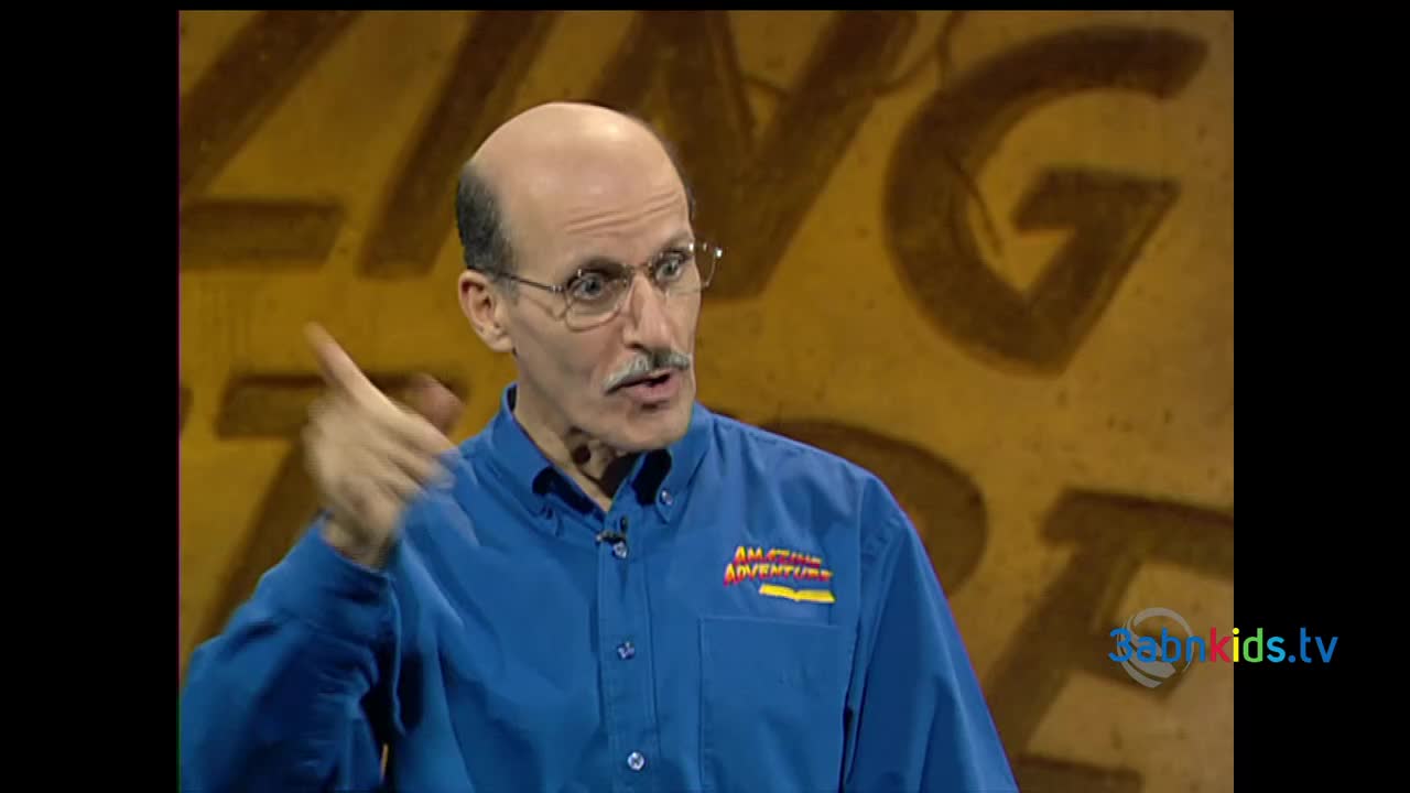 The man in the blue shirt gestures with his right hand, index finger extended. He's speaking on a set with a painted backdrop, presumably for a children's program.
