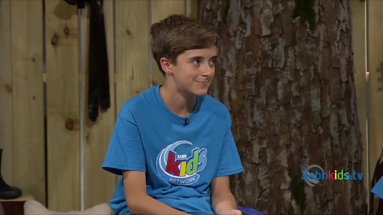 A young man in a blue 3ABN Kids shirt sits with a slight smile, looking off to the side. Behind him, a large tree trunk and wooden planks create a rustic backdrop.
