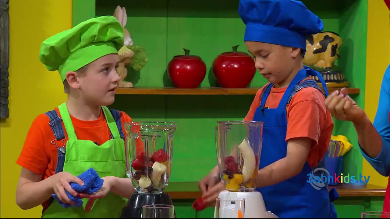 Two young cooks, wearing aprons and chef hats, are adding fruit to blenders. One boy holds a blue cloth, while the other carefully places a strawberry into his blender.
