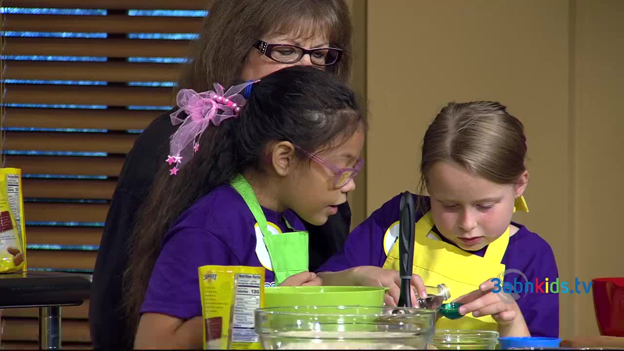 Two young girls, wearing aprons, carefully measure ingredients into a bowl, guided by a woman wearing glasses. The scene is part of a cooking segment on 3ABN Kids, a children's program from the United States.

