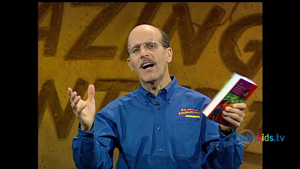 A man in a blue shirt gestures with his left hand while holding a book in his right. The 3ABN Kids logo is visible on his shirt, and the background has the word "AMAZING" painted on it.
