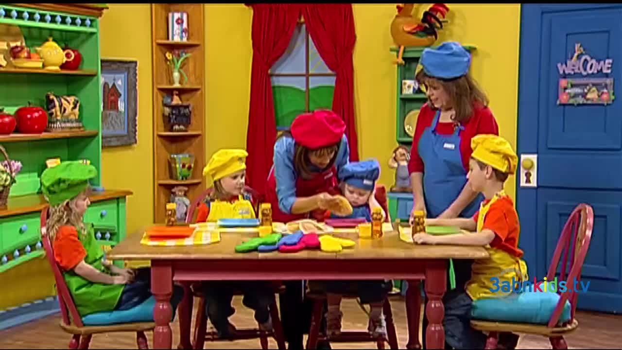 A woman in a red beret is showing a young child how to work with dough, while other children in chef's hats sit at the table. The scene is a brightly-colored set, likely for a 3ABN Kids show.
