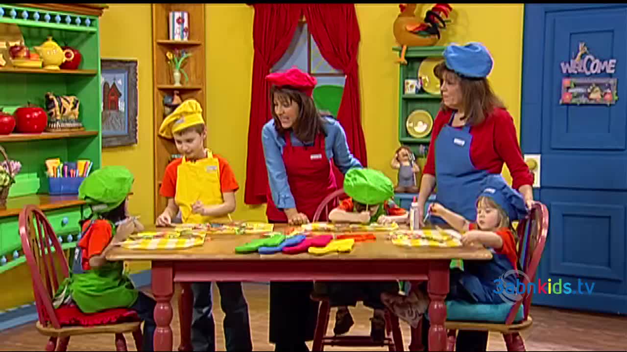 Children in chef hats and aprons sit around a table, working on crafts with the help of two adults. The scene is from a 3ABN Kids program, filmed in a colorful kitchen.
