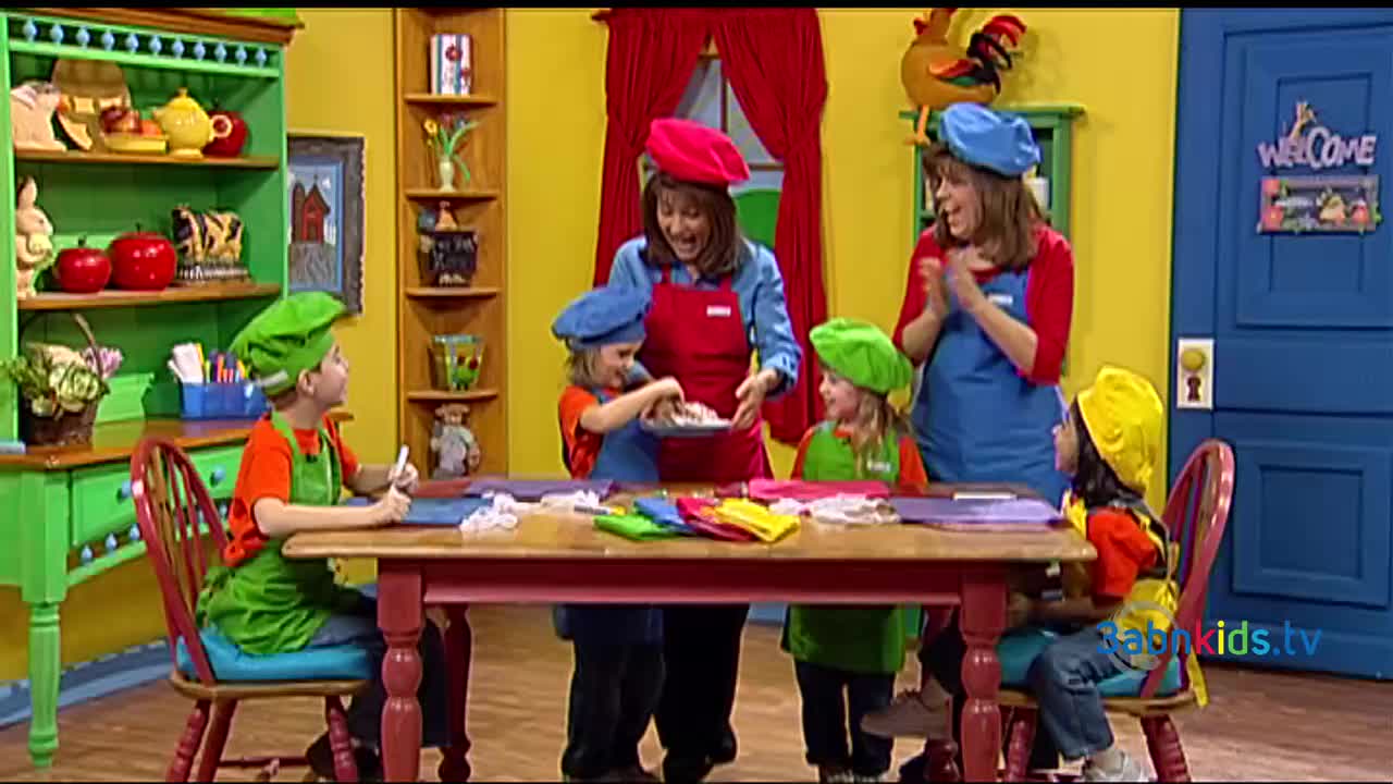 A group of children and adults in aprons and chef hats gather around a table, some are working with materials, while others are holding a bowl. One woman claps as the children work on a craft project for 3ABN Kids.

