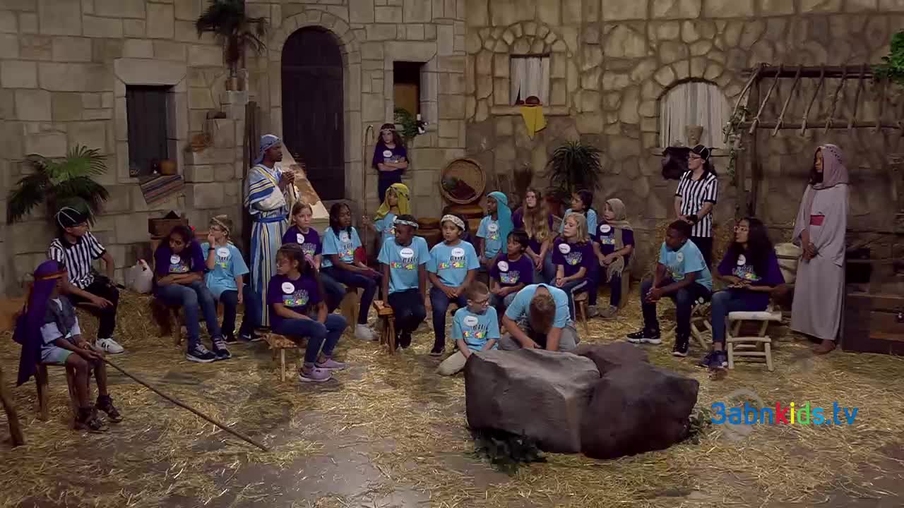 A group of kids in matching shirts are seated on benches, watching a performance on a stage set up to look like an ancient village. A few people in costumes stand nearby, including a person dressed in a referee's uniform.
