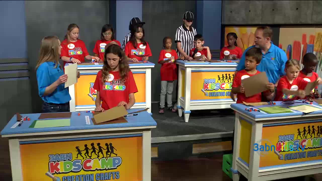Kids in red t-shirts, part of a 3ABN Kids Camp, are busy at their tables working on crafts. The adults, dressed in blue, are assisting them, and a referee in black and white stripes is watching.
