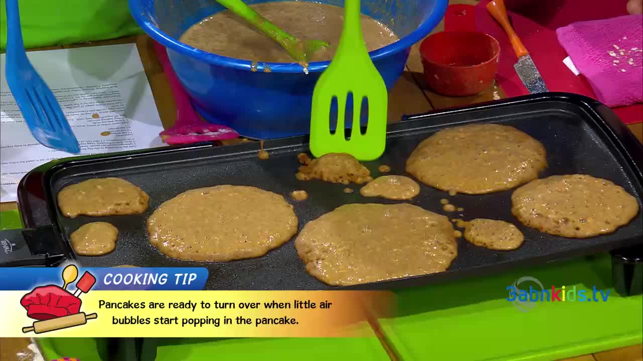 Pancakes are cooking on a griddle, and small bubbles are forming on their surfaces. A green spatula sits ready, and a cooking tip graphic gives instructions.
