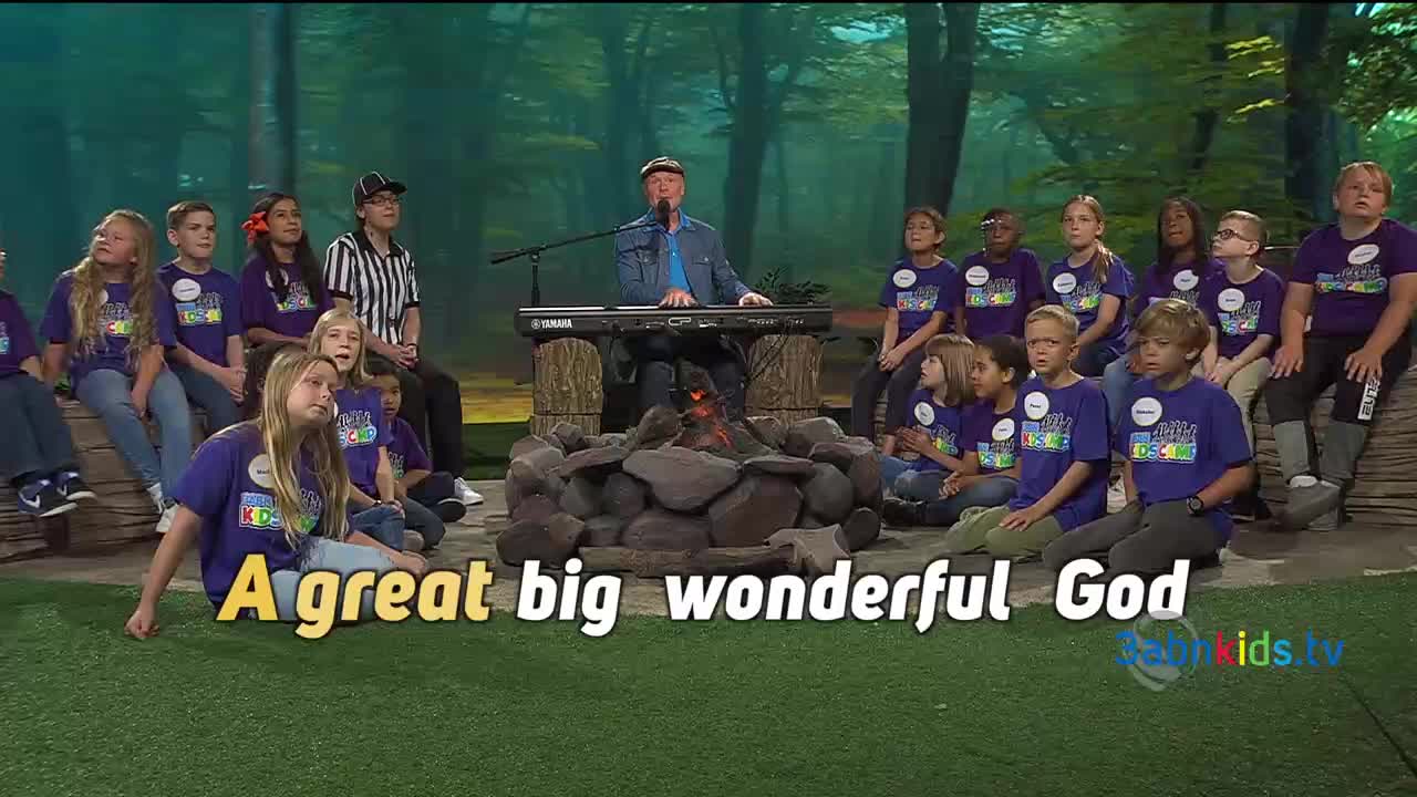 A man plays a keyboard as children in matching shirts sit around a fake campfire. The words "A great big wonderful God" are superimposed on the grass in front of them.
