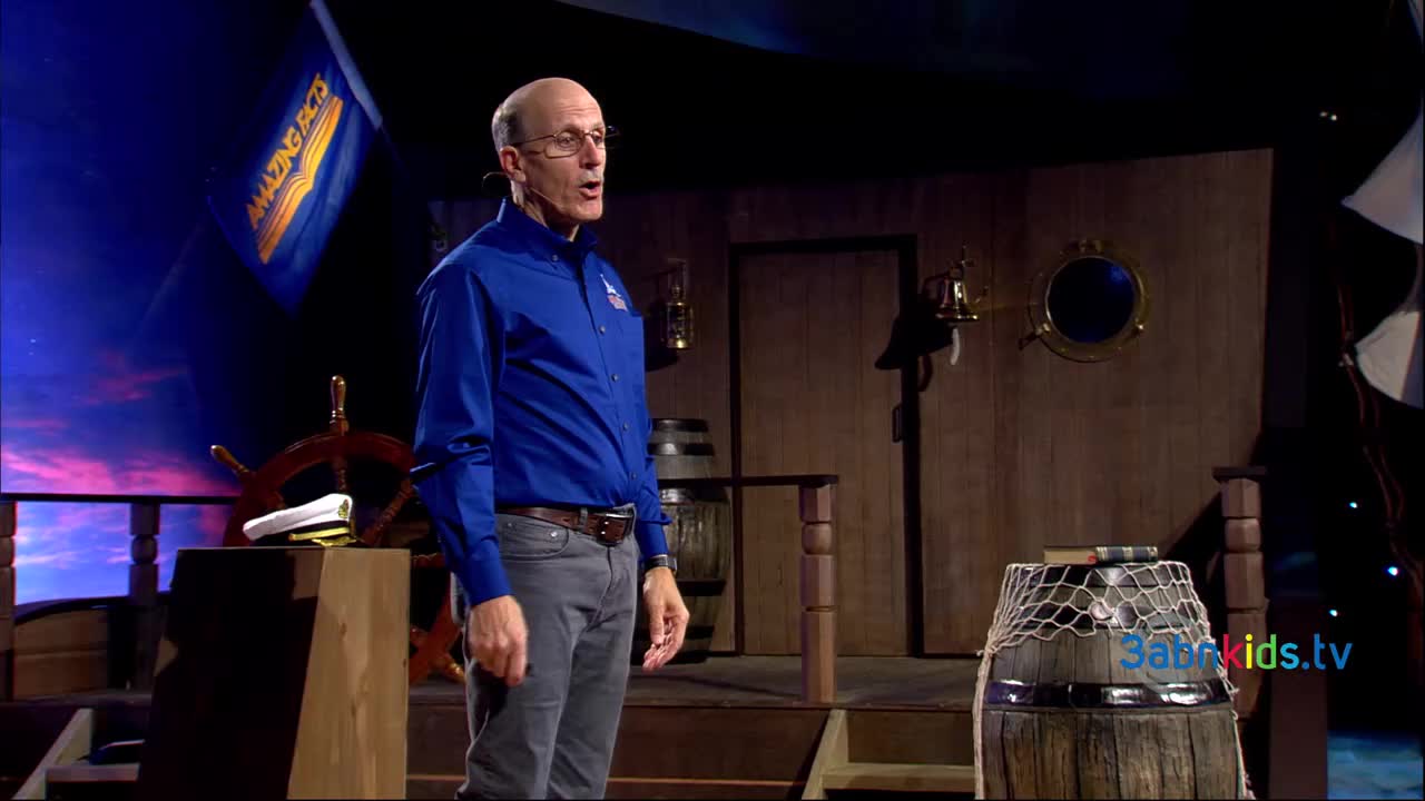 A man in a blue shirt and glasses is speaking on a stage designed like a ship's deck, with a "Amazing Facts" flag in the background. He's gesturing with his hands, likely addressing the audience of 3ABN Kids.
