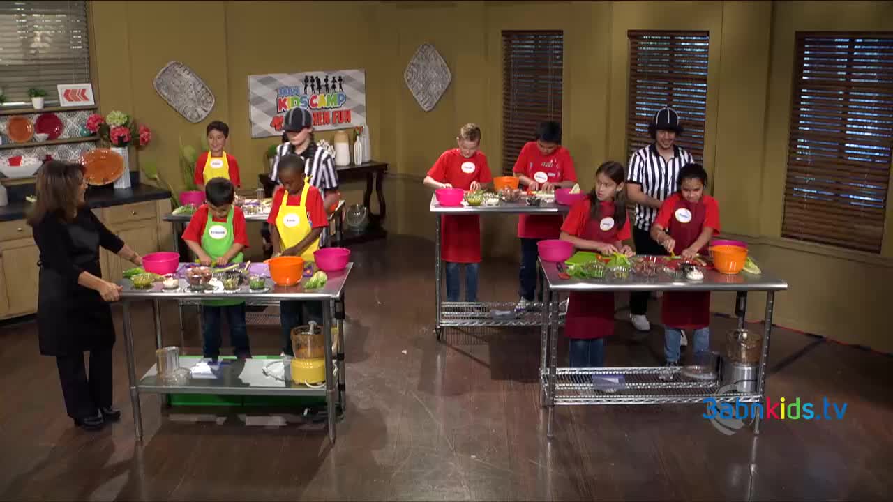 Kids in red shirts and aprons are focused on chopping vegetables at stainless steel tables. A woman in black oversees the activity, and a referee-looking person watches the kids.
