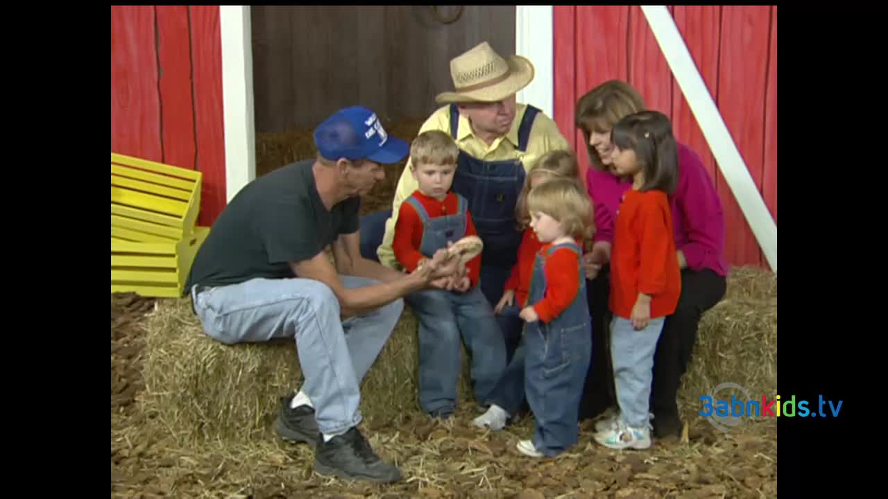 A man in a blue cap and another in a straw hat are showing a small group of children something held between their hands. The kids, dressed in overalls and red shirts, lean in to get a better look.
