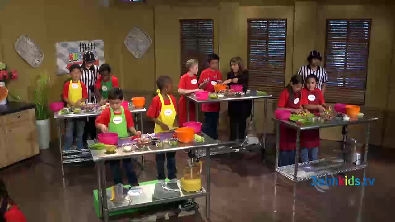 Children in aprons and red shirts are gathered around stainless steel tables, preparing food. A woman in a black jacket is speaking with some of the kids as they work.

