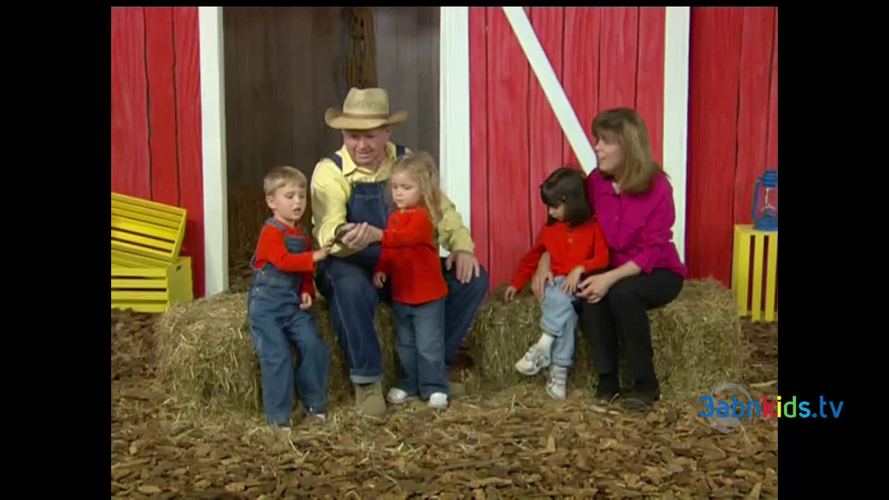 A man in a straw hat sits on a bale of hay, holding something small. Two children in matching red shirts and overalls are watching intently.
