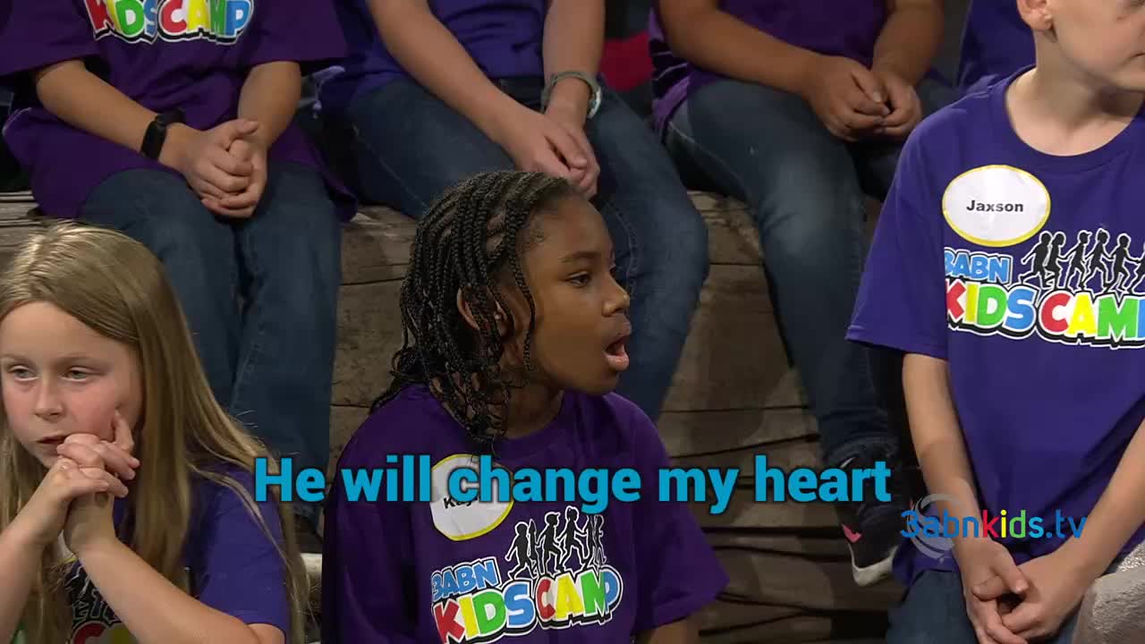 A young girl with braids sings with her mouth open, eyes focused ahead. Other children, wearing matching purple shirts, sit around her, some with their hands clasped.
