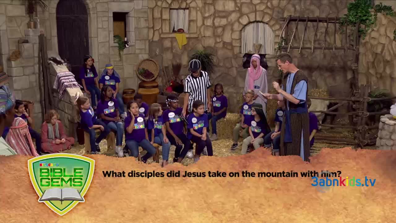 A man in a tunic gestures as he speaks to a group of children, some wearing purple shirts, who are seated on hay bales. The set is designed to resemble a stone structure, and a person dressed in a referee's uniform stands nearby.
