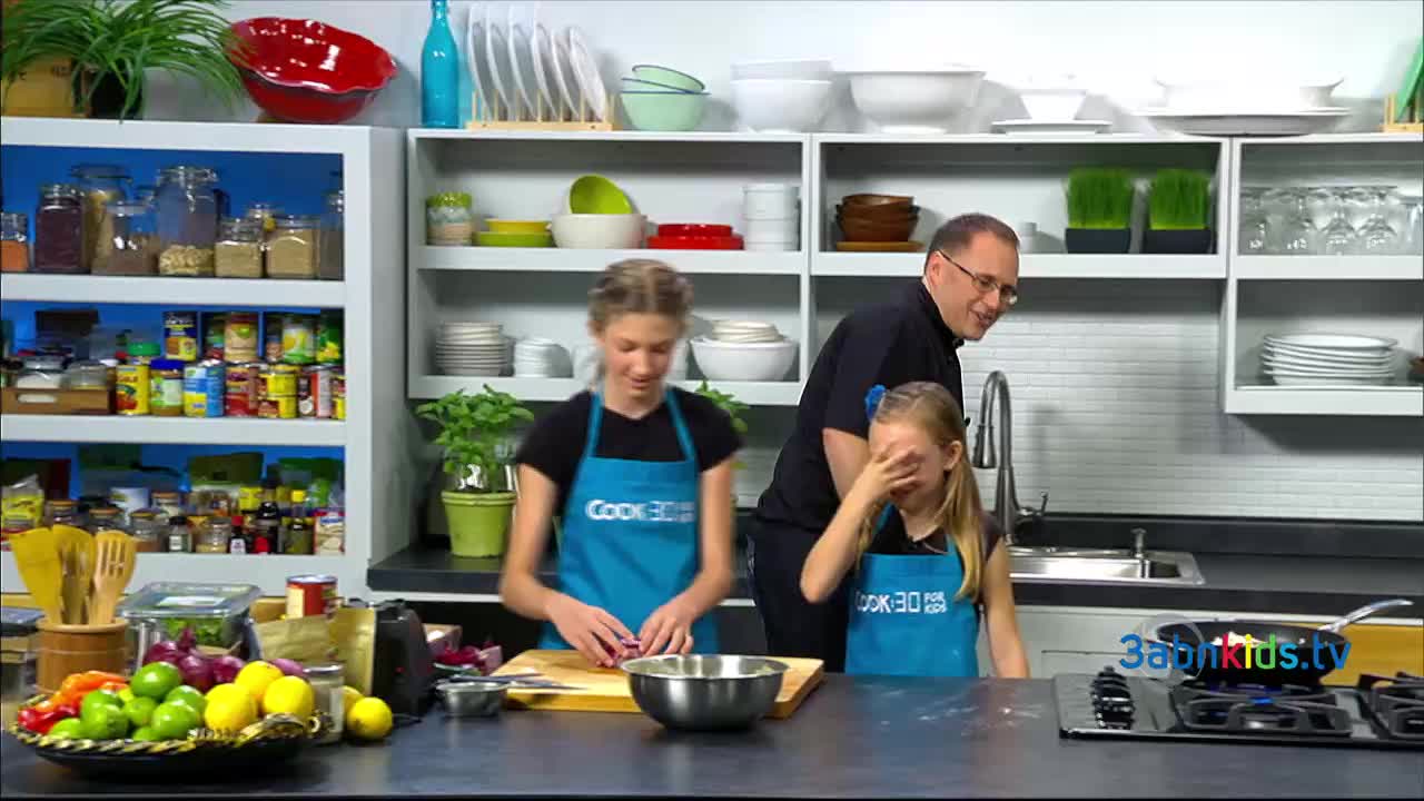 A girl in a blue apron is chopping something on a wooden board, while another covers her face. A man in glasses smiles at the scene, standing near the sink.
