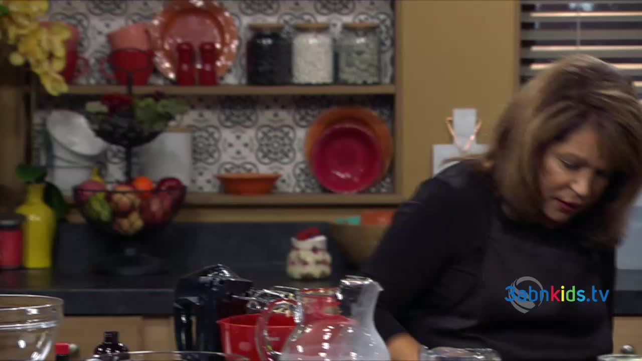 A woman in a black apron leans over a countertop, likely in a kitchen from the United States, on the set of 3ABN Kids. A hand mixer and glass pitcher are visible in front of her.
