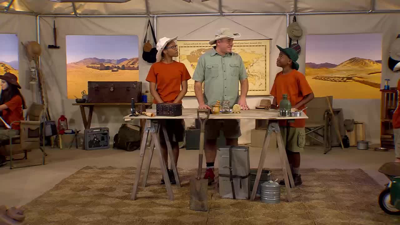 A man in a khaki shirt stands at a makeshift table, talking to two children wearing orange shirts and safari hats. The set is decorated to look like a desert camp, likely for a 3ABN Kids program.
