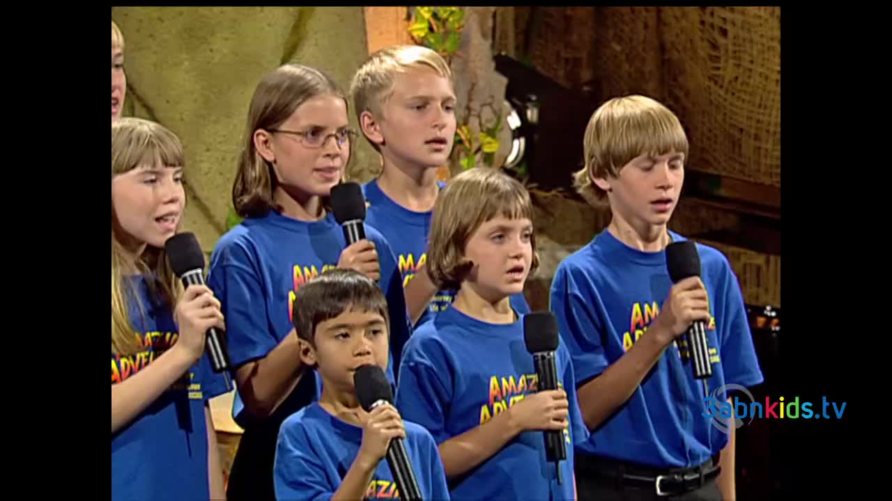 A group of children, all wearing matching blue shirts, are singing into microphones on a stage. The backdrop looks like a jungle scene, and the children's faces show concentration as they perform.
