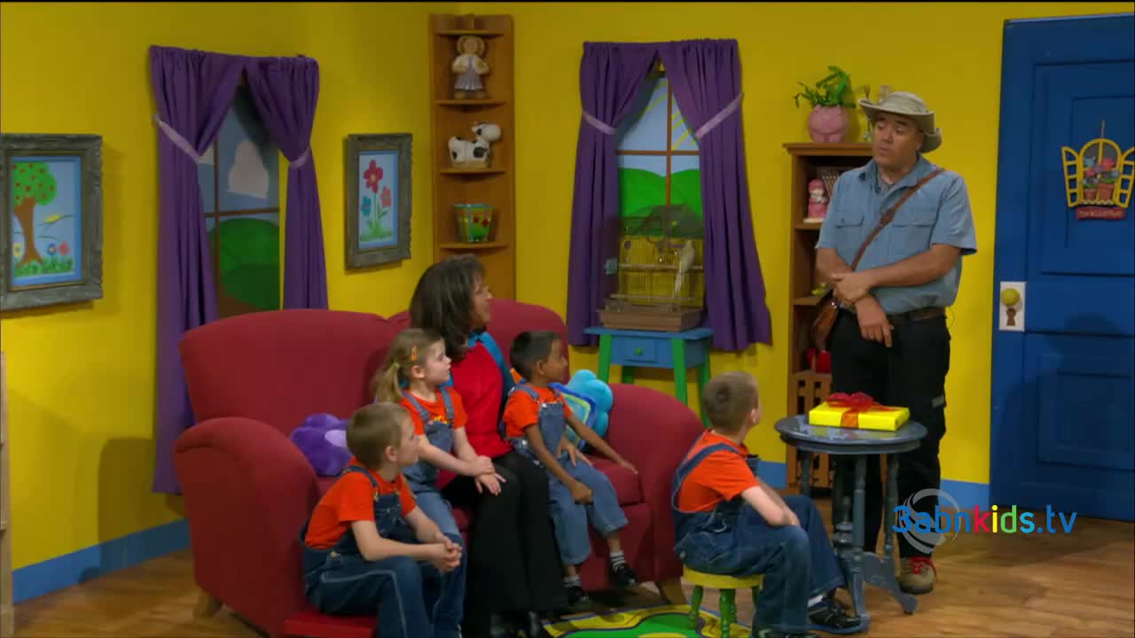 A man in a hat stands beside a small table holding a wrapped gift.  Children in orange shirts and blue overalls sit on a red couch, watching.
