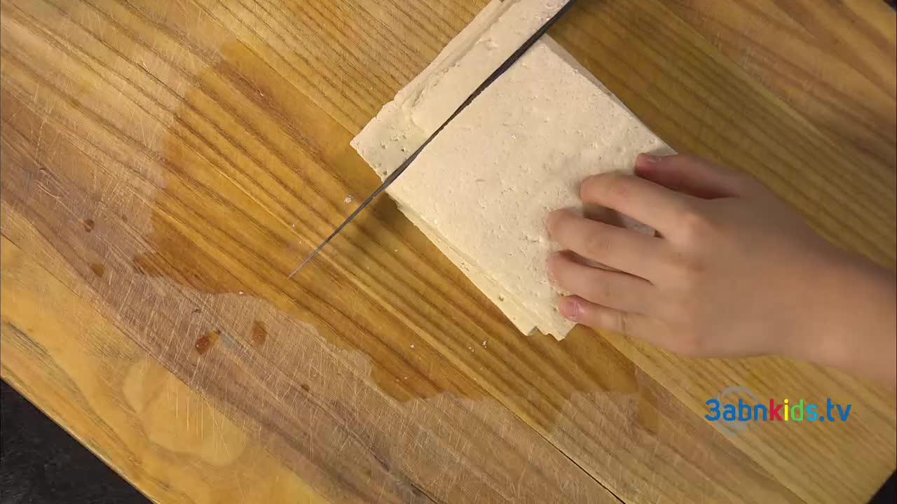 A child's hand carefully guides a knife through a stack of tofu, neatly slicing it into layers. The scene is taking place on a wooden cutting board, likely as part of a cooking segment from 3ABN Kids in the United States.
