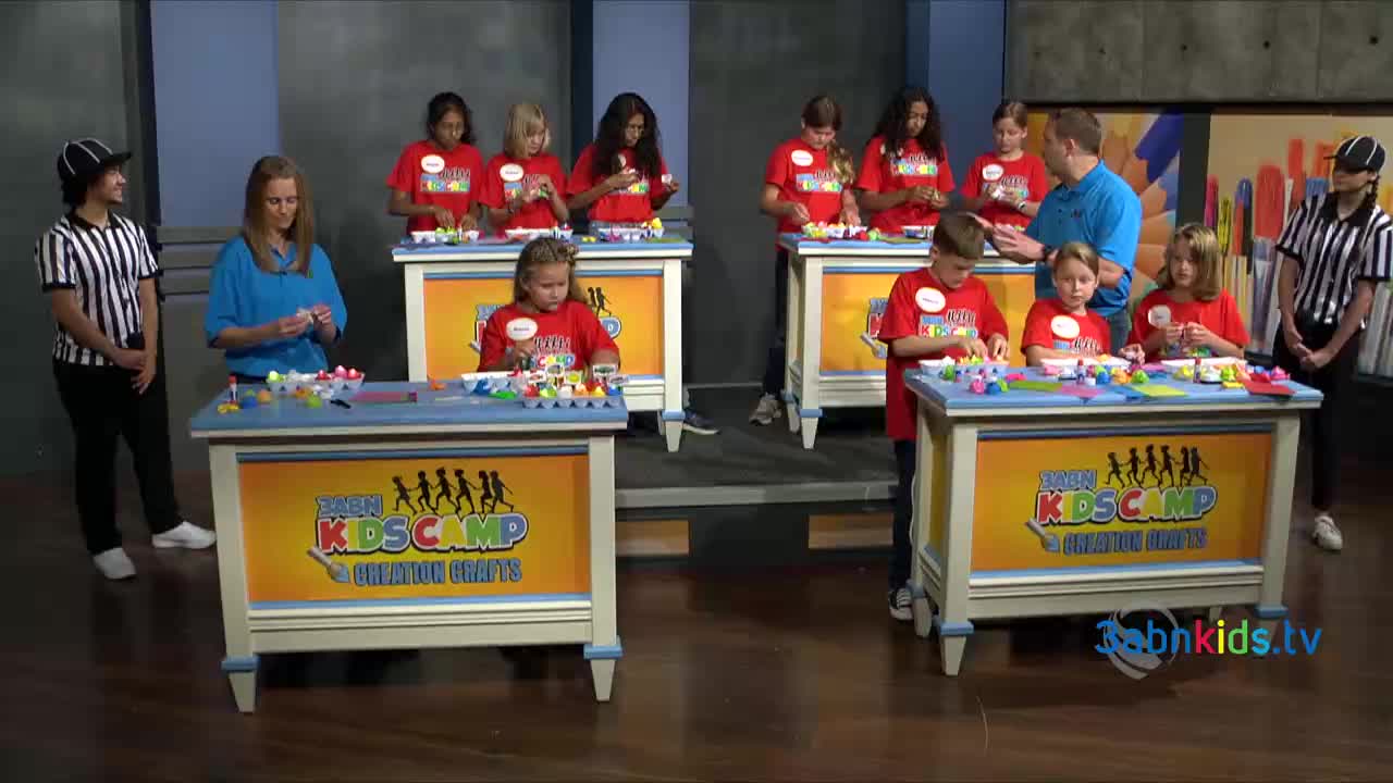 Children in red shirts are focused on crafting at individual tables, while adults and referees observe. The scene takes place on a 3ABN Kids set, with the logo visible on the tables.
