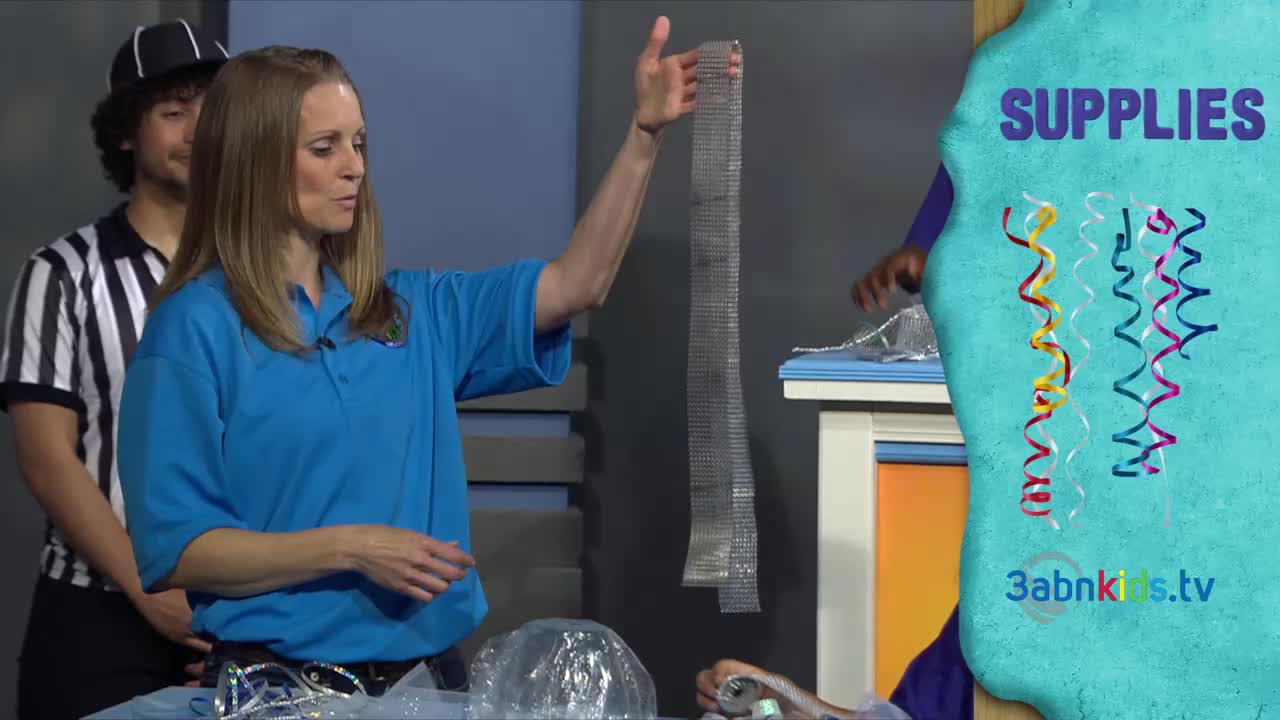 A woman in a blue shirt holds up a long, silver mesh strip for the camera. Beside her, a man in a referee uniform and cap looks on as the 3ABN Kids show gets underway.
