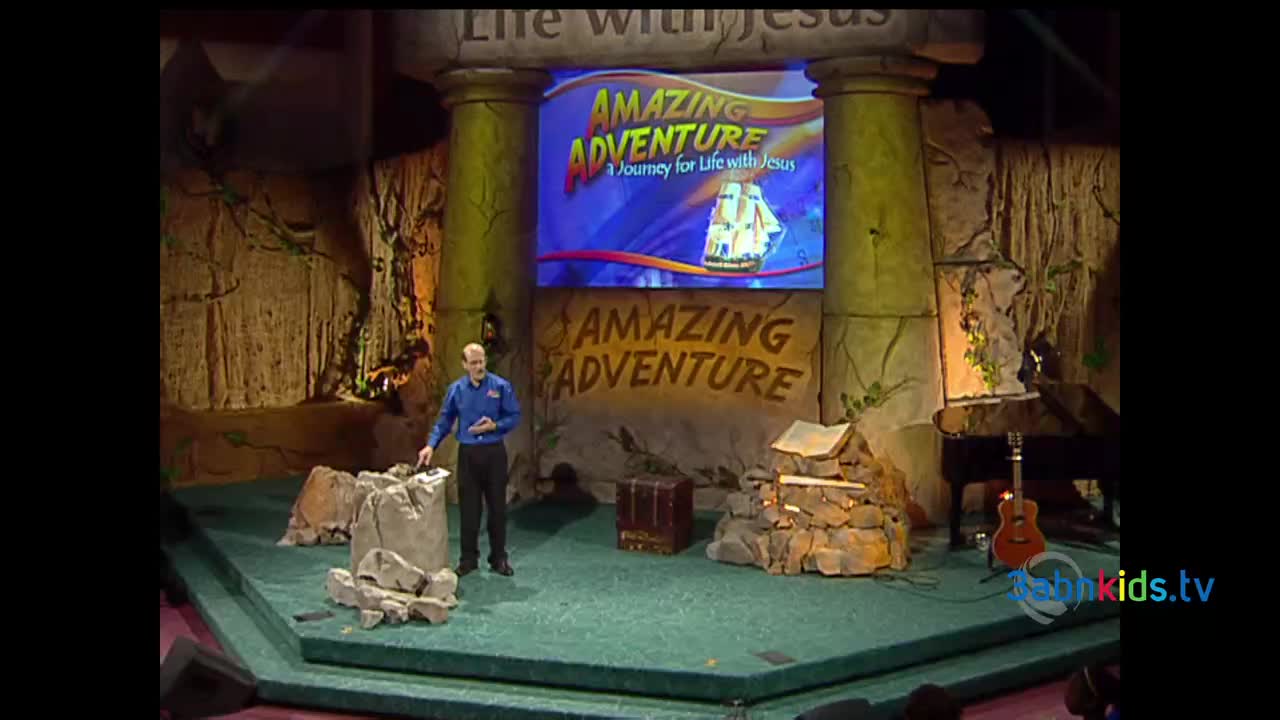 A man in a blue shirt gestures toward a pile of faux rocks on a stage set. Behind him, a screen displays the words "Amazing Adventure" and a picture of a sailing ship, likely for a 3ABN Kids program.
