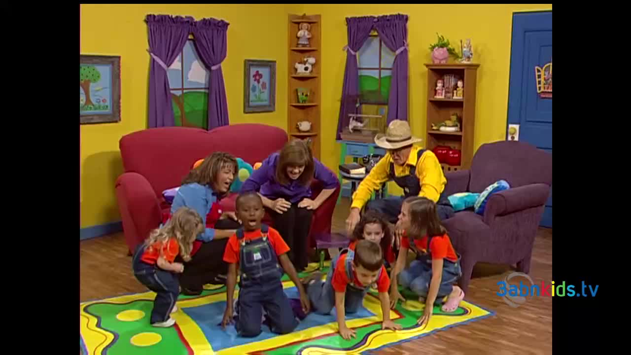 A group of children are crawling on a colorful rug, while adults watch and encourage them. The scene is set in a brightly decorated room, likely for a children's show in the United States.
