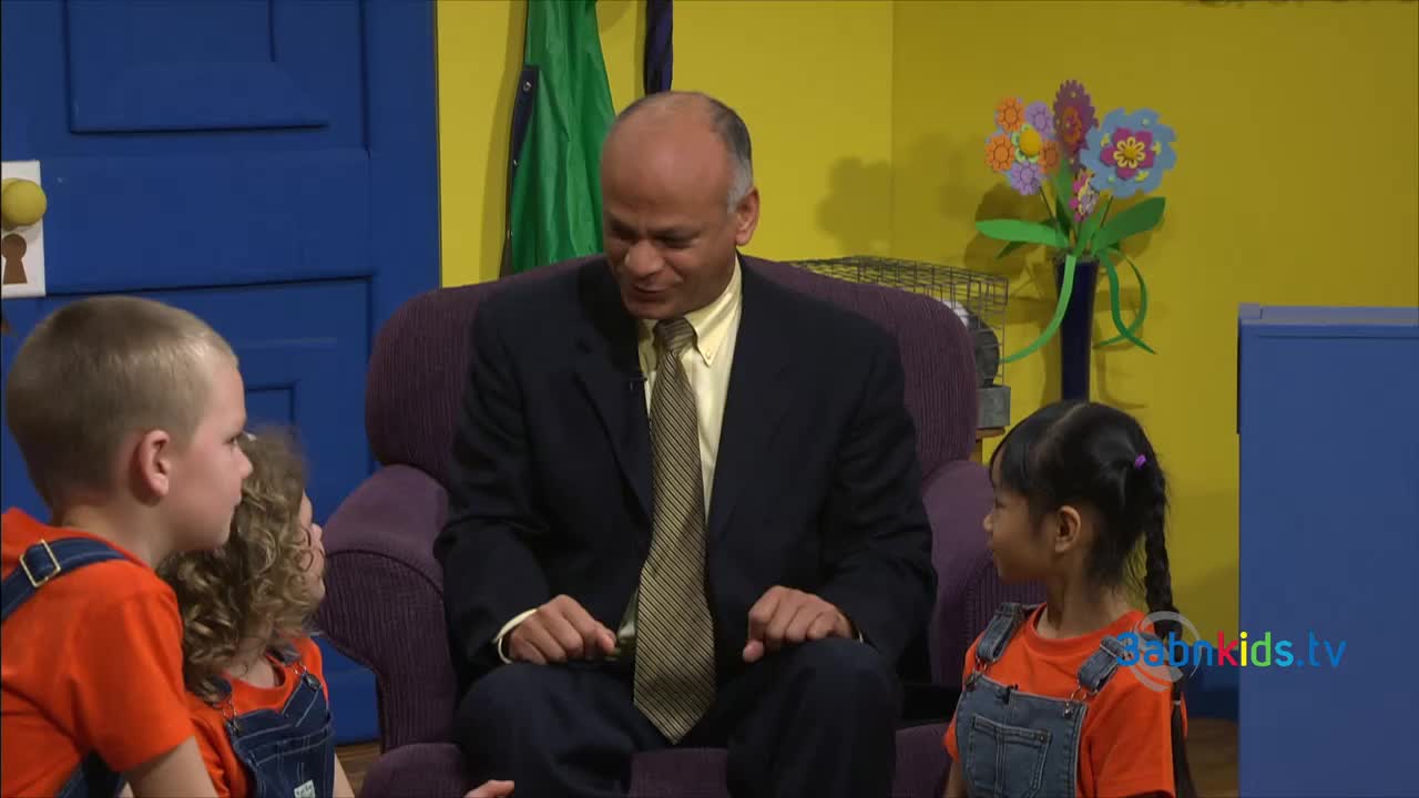 A man in a suit sits in a purple armchair, talking to three children in orange shirts and overalls. The set has a bright, colorful backdrop with a blue door and a yellow wall.
