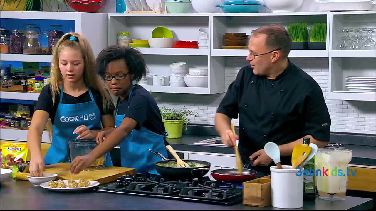 Two young women, wearing aprons, are carefully arranging golden-brown food items on a plate. A man in a chef's coat stirs something in a frying pan, as the scene unfolds in a kitchen setting.
