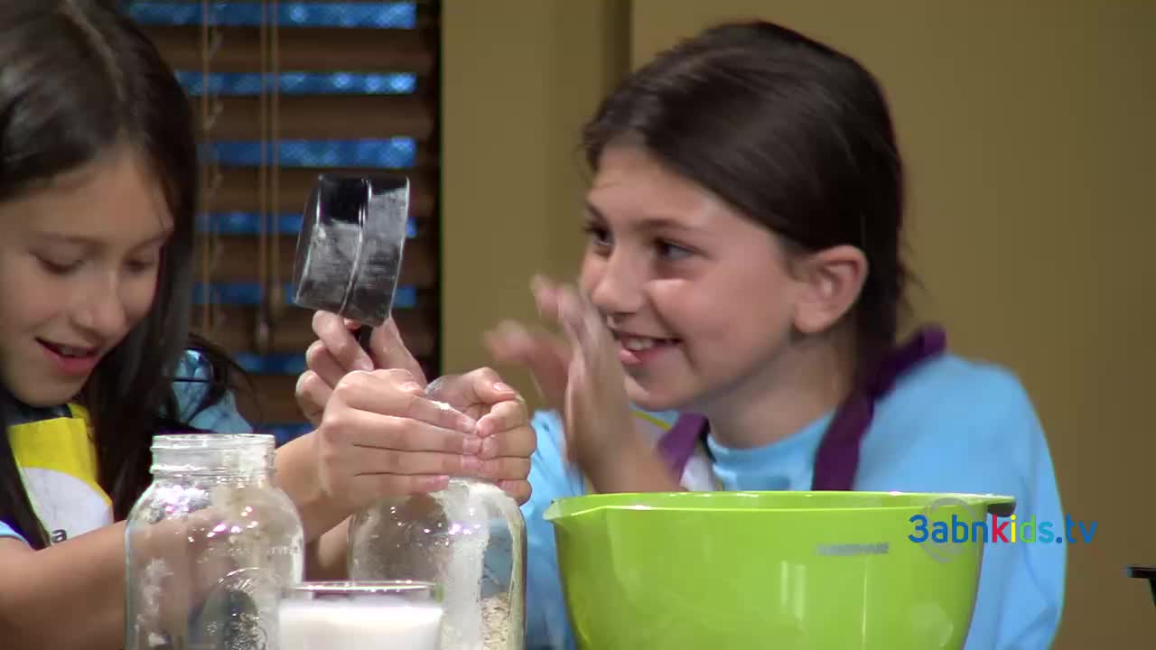 Two girls are in a kitchen, one measuring something into a jar with a metal scoop. The other girl is smiling, seemingly delighted by the activity.
