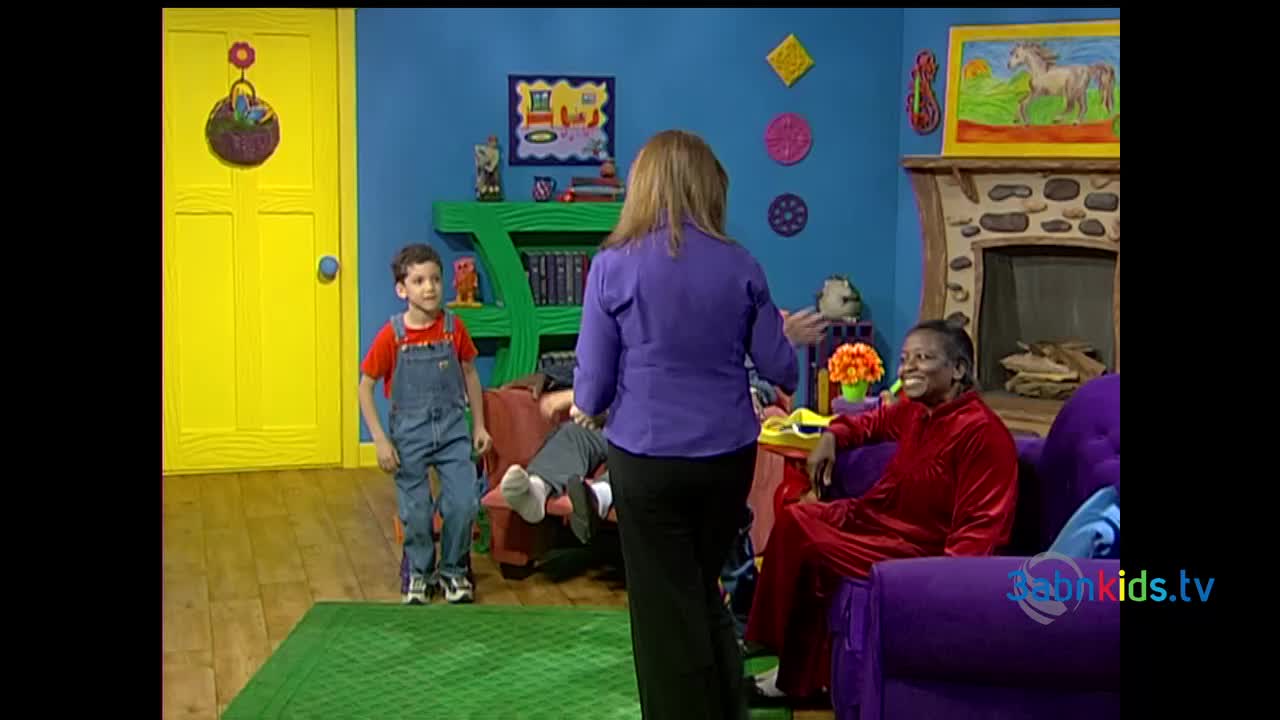 A woman in a purple shirt gestures toward the boy in overalls, while another person relaxes on a sofa. The set, with its bright colors and cartoonish decorations, looks like it's from a 3ABN Kids show.
