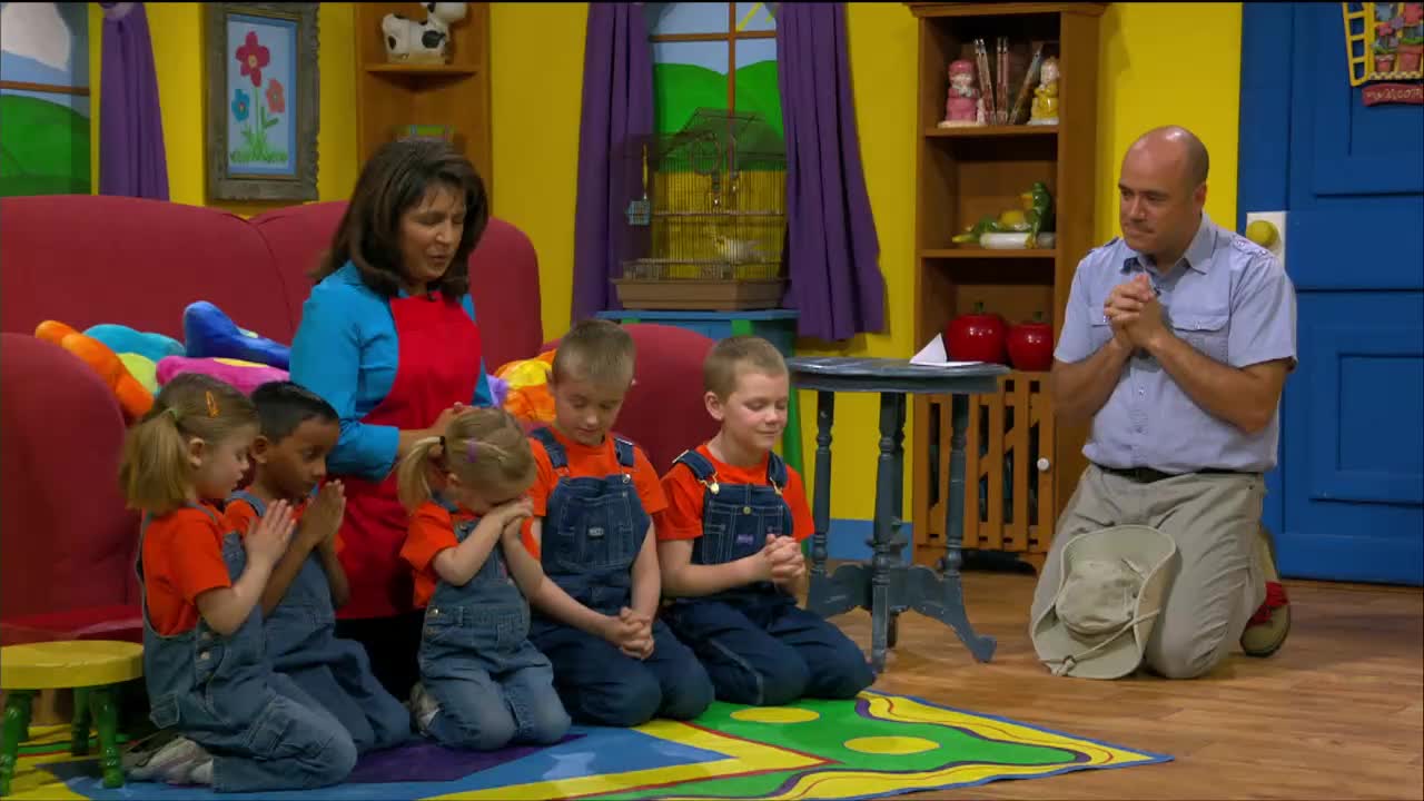 A group of children and two adults are kneeling, hands clasped in front of them. They're all bowing their heads, as if in prayer, in a brightly colored room.
