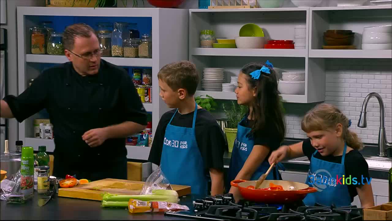 A chef in a black coat gestures towards a cutting board as three children in aprons watch. One girl stirs vegetables in a red pan on the stove.
