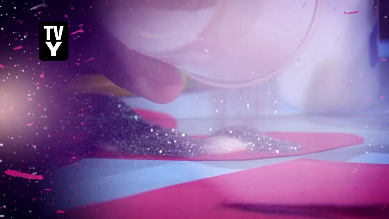 A hand tips a clear container, and a stream of silver glitter falls onto pink and blue paper. Tiny particles float in mid-air, catching the light.
