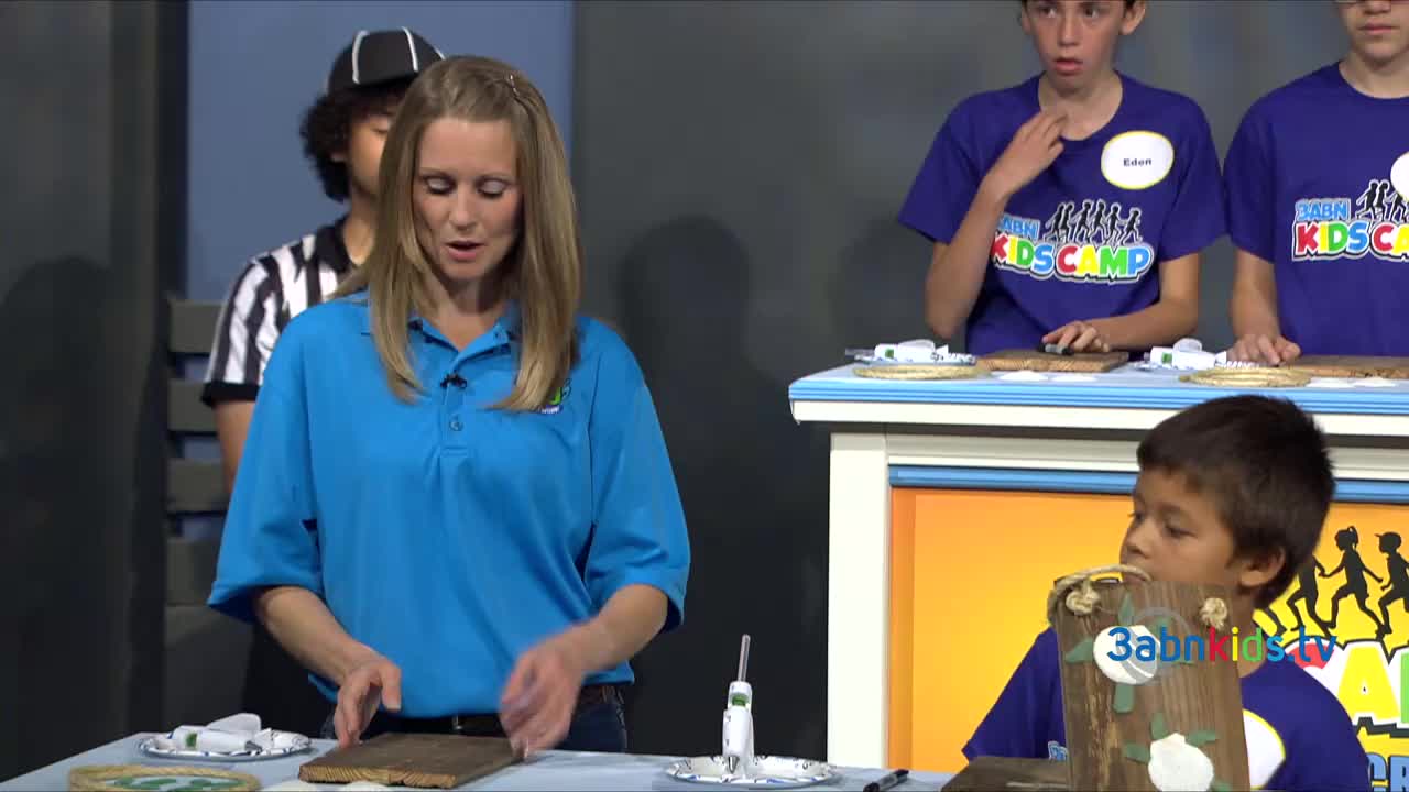 A woman in a blue shirt gestures toward a wooden board on the table. Across the room, children in matching shirts are focused on their own projects.
