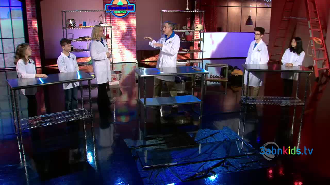 A man in a lab coat gestures toward a stainless steel table, while a group of kids in similar coats stand at other tables. The set, lit with bright colors, appears to be for a science show.
