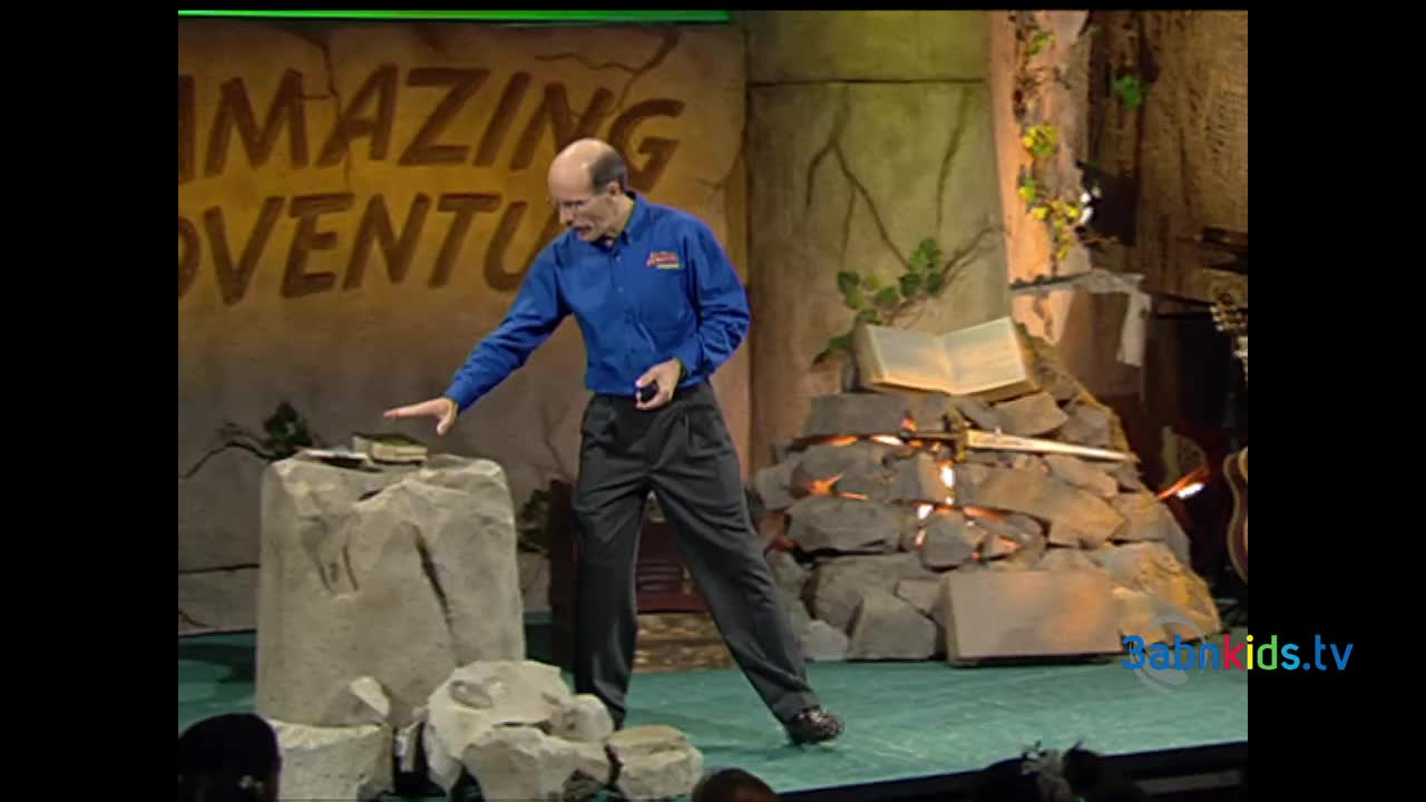 A man in a blue shirt gestures toward a book resting on a stone. Behind him, a backdrop reads "Amazing Adventure," likely part of a 3ABN Kids production.
