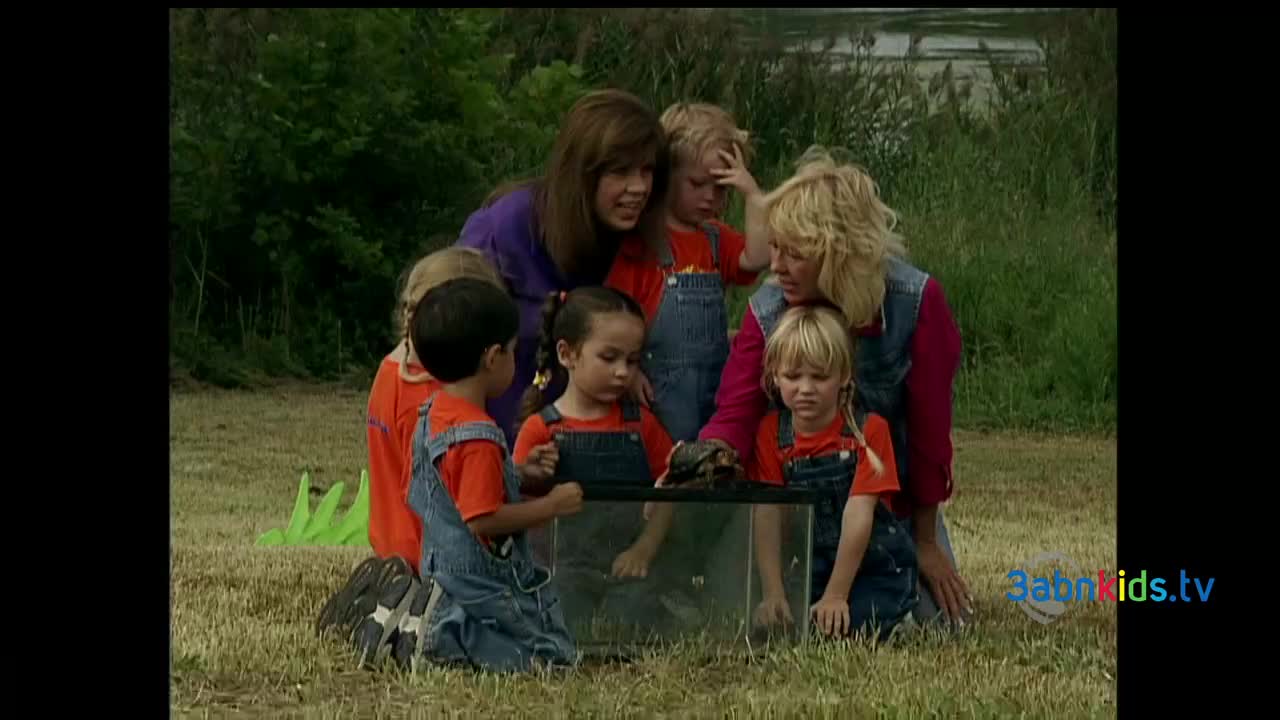 A group of children in orange shirts and denim overalls are gathered around a clear container. Inside, a turtle sits while the kids and two women watch intently.
