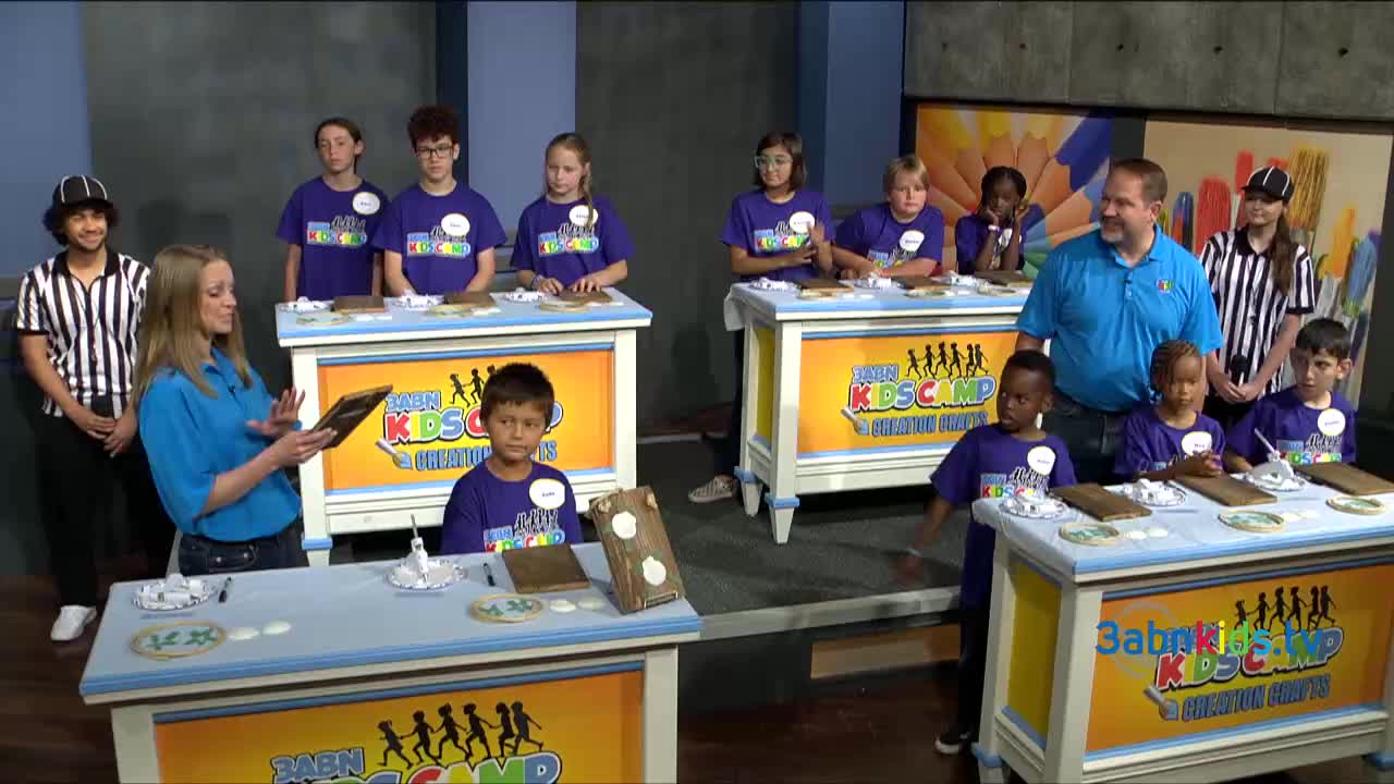 A woman in a blue shirt holds a tablet, addressing a group of children. The kids, wearing matching purple shirts, are at tables with various items in front of them.
