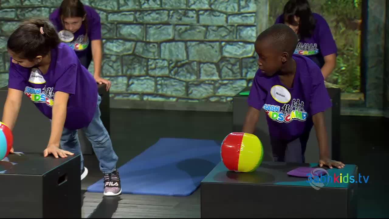 Children in matching purple shirts are leaning on black cubes, each with a colorful ball nearby.  They appear to be part of a 3ABN Kids activity, with a blue mat and a brick wall backdrop.
