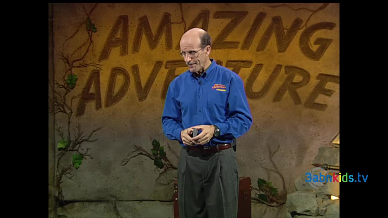 A man in a blue shirt and glasses stands in front of a backdrop that says "Amazing Adventure." He's holding something small in his hands, looking down.
