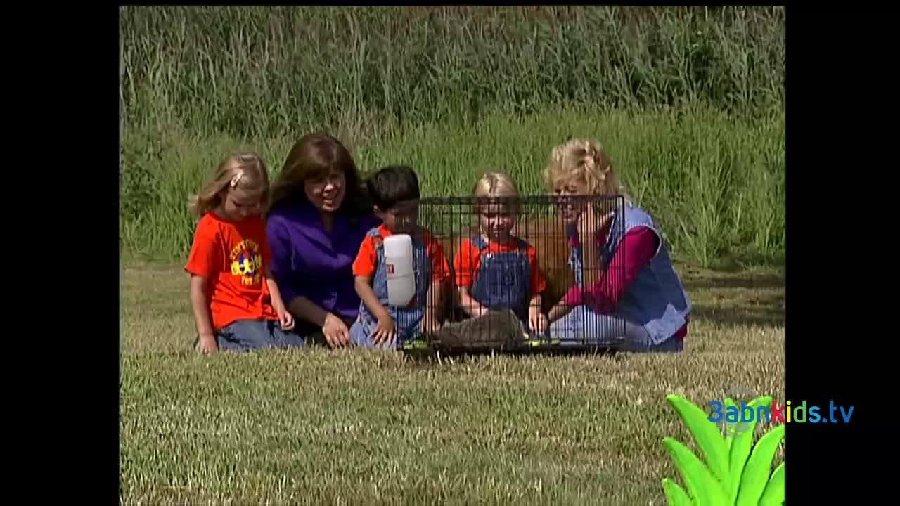 A group of children and adults are gathered around a small cage on the grass. Inside, a small, furry animal is nestled, while the kids watch with interest.
