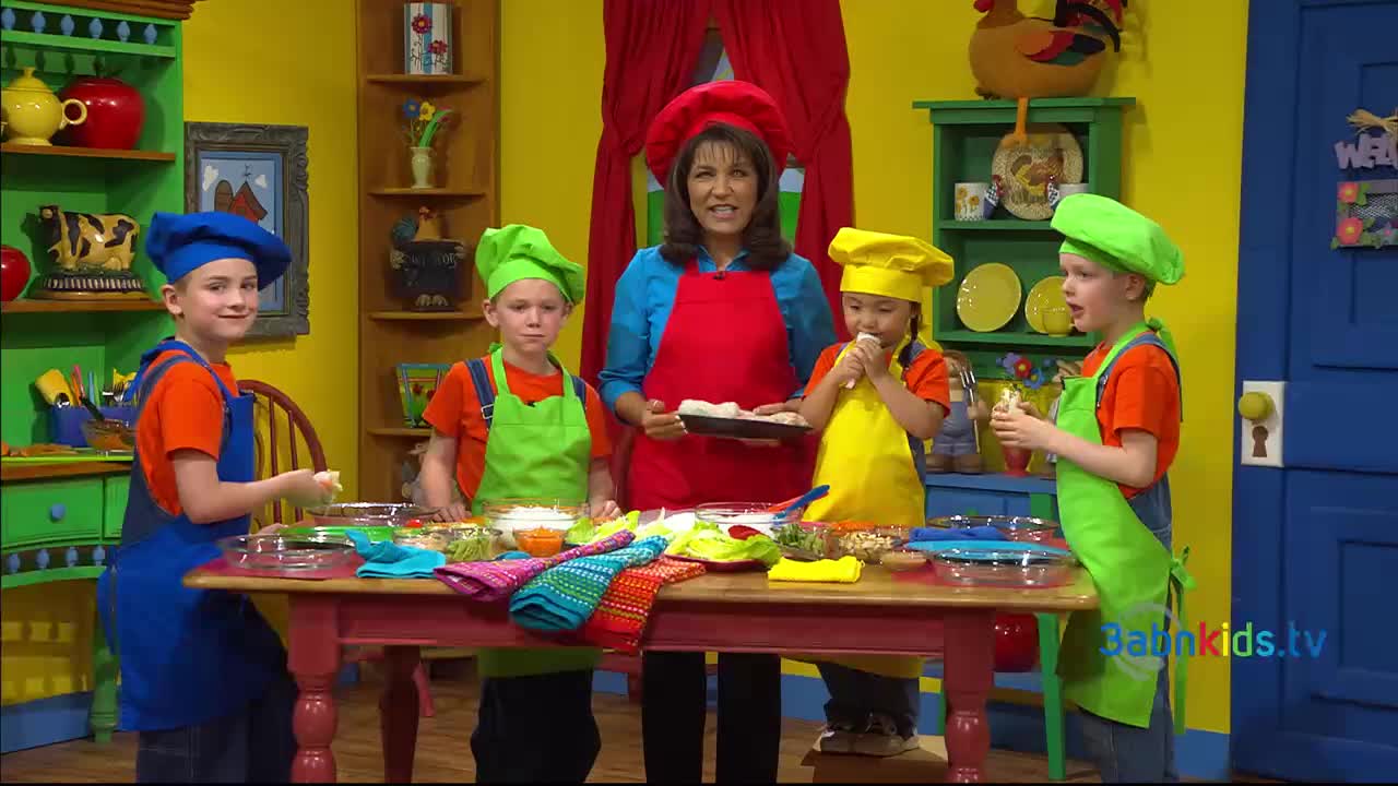 A woman in a red apron holds a tray of something while the children around the table, all in chef hats and aprons, prepare food. One of the kids is enjoying a snack, and the scene looks like it's from a 3ABN Kids show.
