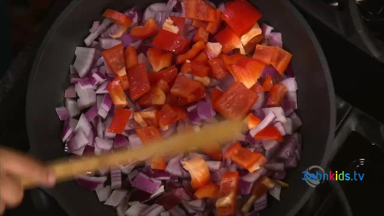 Diced red bell peppers and purple onions sizzle in a pan, stirred by a wooden spoon. This is likely a cooking segment on 3ABN Kids, a children's program from the United States.

