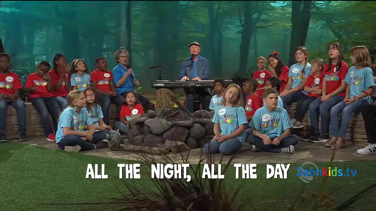 A man plays a keyboard as a group of children, some in red shirts and others in blue, sing along. The scene is set with a backdrop of trees and a stone fire pit in front.
