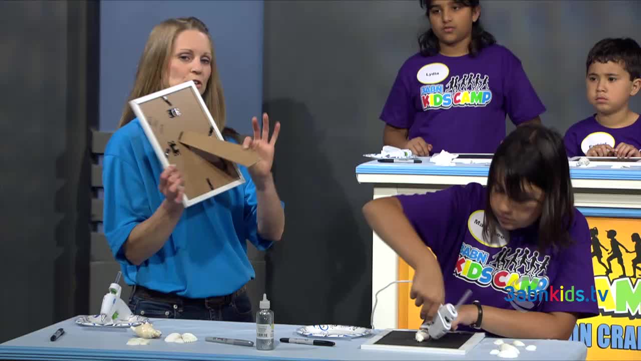 A woman in a blue shirt holds a picture frame, showing the back.  A girl in a purple shirt uses a glue gun on a black surface, surrounded by seashells.
