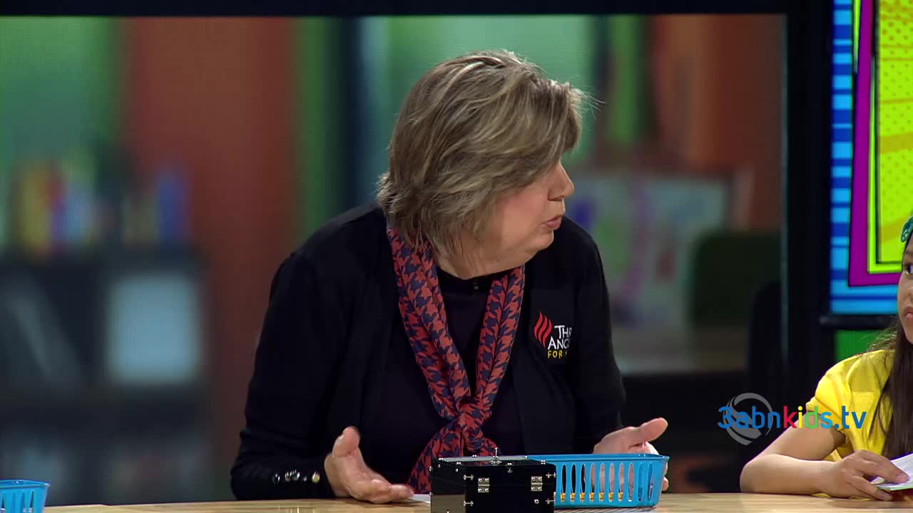 The woman in the black cardigan gestures with her hands as she speaks. A young girl in a yellow shirt sits beside her, listening attentively on the set of 3ABN Kids.
