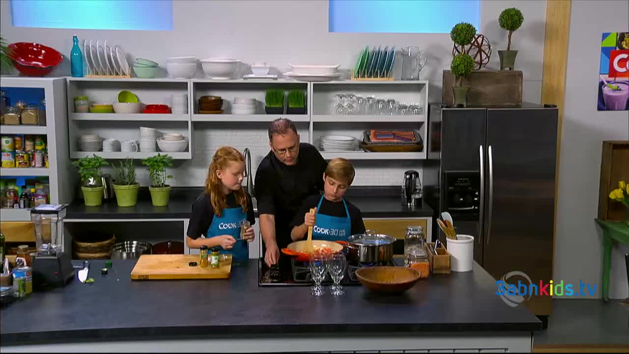 A man and two kids, all wearing aprons, are gathered around a stovetop, cooking. The boy stirs a pot with a wooden spoon while the girl sprinkles something from a small container.
